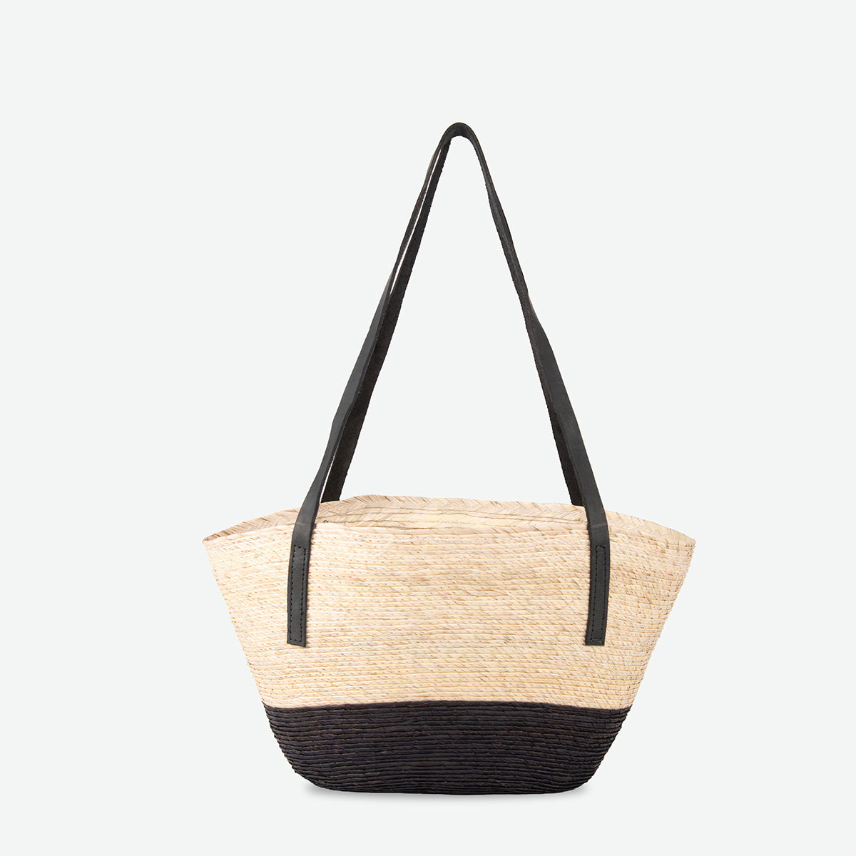 A woven straw tote bag with a natural beige body and a black bottom base, featuring two long black shoulder straps, shown against a plain light gray background.