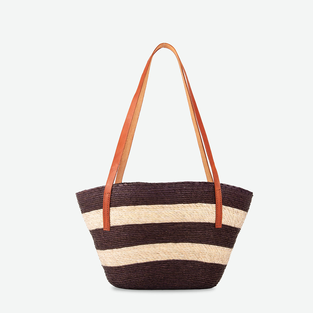 A woven straw tote bag with horizontal dark brown and beige stripes, featuring two long tan leather shoulder straps, shown against a plain light gray background.